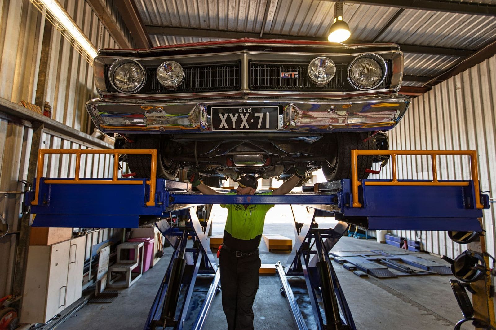 Mechanic inspecting the underside of a classic car raised on a workshop hoist.